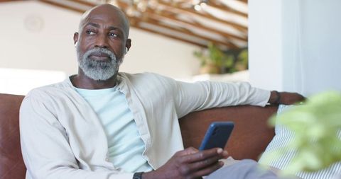 Senior african american man sitting on leather sofa holding smartphone, looking thoughtful