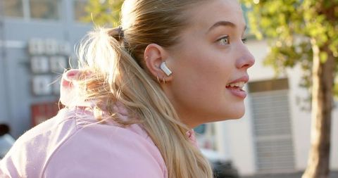 Young woman leaning forward listening with wireless earbuds during golden hour on street