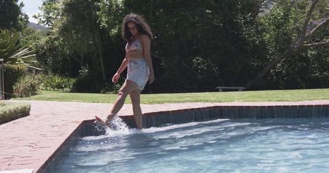African woman splashing poolside in bikini and sunglasses on sunny backyard patio