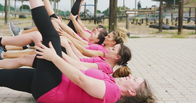 Group of women wearing pink shirts participating in outdoor fitness class, performing leg raises while lying on ground. Useful for promoting group fitness activities, health and wellness events, and outdoor exercise programs. Highlights teamwork and active lifestyle.