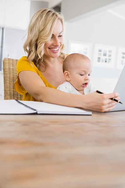 This image of a mother working on her laptop with her baby on her lap in a home kitchen setting is ideal for articles or advertisements about work-from-home, multitasking parents, parental advice, family life balance, and child care. It can also be used in blogs or websites that discuss remote working, parenting tips, and family routines.