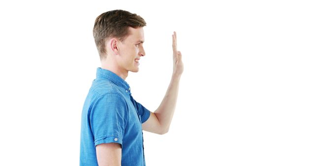This image features a young man in a blue shirt waving his hand against a white background. He stands casually and is viewed from the side. This photo can be used in contexts such as social interaction, online communication, presentations, advertisements, and websites to depict greeting, friendliness, or farewell.