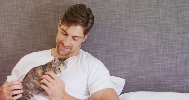 Man wearing white shirt lounging on bed with a kitten in relaxing home environment. Could be used for content related to pet care, relaxation at home, or human-animal bonding.
