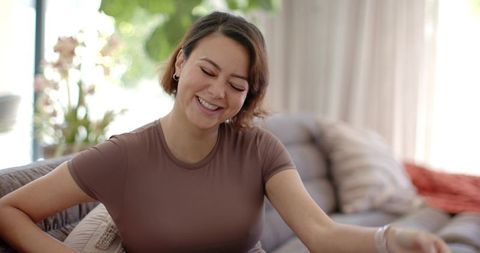 Smiling woman relaxing on gray sofa reaching forward in sunlit living room with plants