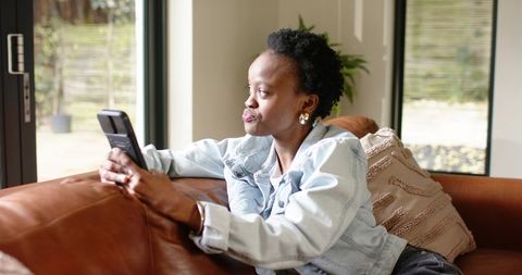 Contemplative african american woman sitting on sofa holding smartphone in cozy room