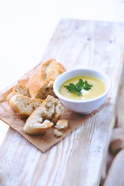 Creamy soup garnished with parsley and served with fresh baguette pieces on a rustic wooden table. Ideal for food blog posts, restaurant menu designs, café advertising, or any visual depiction of comfort food and homemade meals.