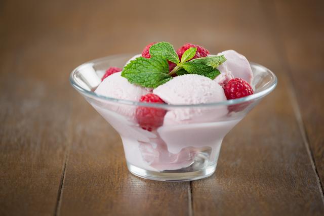 Raspberry ice cream served in a glass cup, garnished with fresh mint leaves and whole raspberries, placed on a wooden table. Ideal for use in food blogs, dessert recipes, summer treat promotions, and gourmet dessert menus.