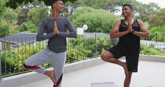 Two men balancing in tree pose during outdoor yoga session on balcony. Ideal for promoting fitness, healthy lifestyle, outdoor exercise, and yoga practice. The greenery in the background emphasizes nature's calming influence. Suitable for health and fitness magazines, wellness blogs, and yoga class advertisements.