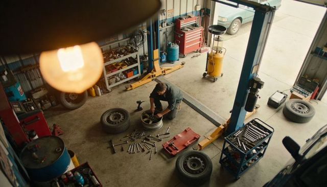 Mechanic kneeling tightening wheel in garage with two-post lift and scattered tools