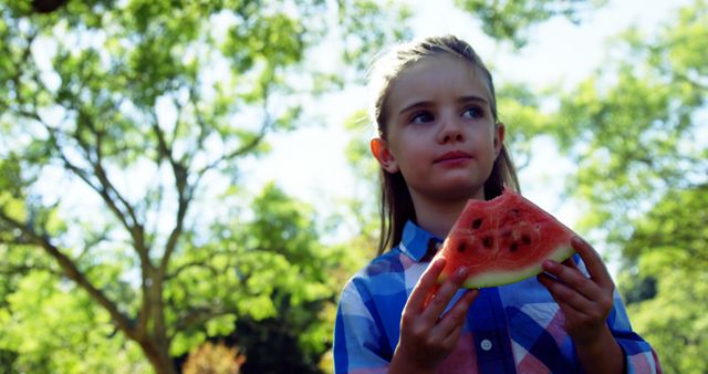 A young Caucasian girl enjoys a slice of watermelon outdoors, with copy space. Her thoughtful expression and the natural setting suggest a moment of summertime leisure.