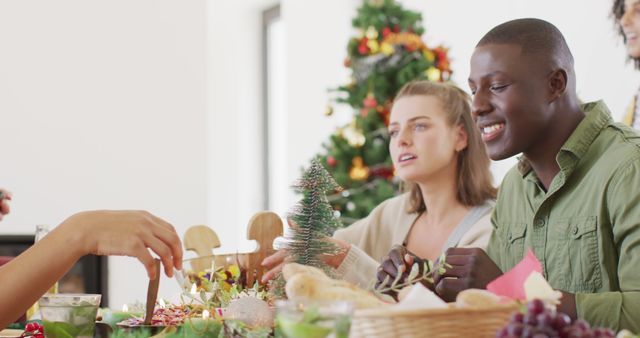 Friends gathered around table sharing Christmas dinner, diverse group bonding over holiday meal. Decorated tree in background creates festive atmosphere. Perfect for holiday greeting cards, festive social media content, promoting holiday gatherings, advertising holiday food products.