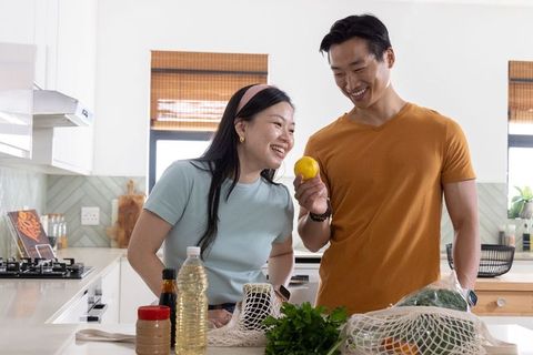 Asian couple preparing groceries and smiling while examining lemon in modern kitchen
