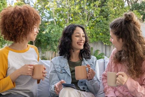 Multicultural women enjoying coffee and chatting on outdoor sofa, candid backyard lifestyle