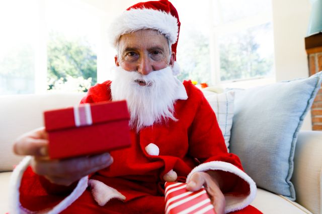 Front view of an old caucasian man wearing a santa clause costume holding out a christmas gift in front of him. he is sitting on the couch in the living room.