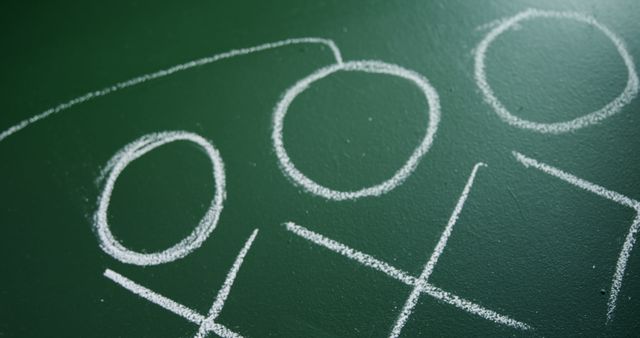 Tic-tac-toe game drawn in chalk on green chalkboard conveys a classroom or educational environment. Suitable for illustrating concepts of strategy and brain games, especially in education, tutorials, and student activities.