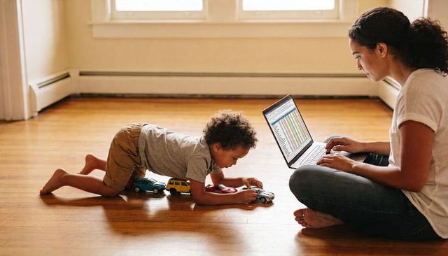 Working parent using laptop while child playing with toy cars on hardwood floor