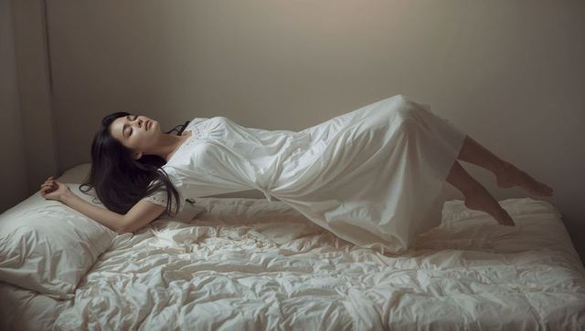Asian woman levitating above bed in white nightgown, dreamy minimalist bedroom with soft light