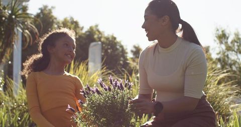 Diverse mother and daughter tending lavender, smiling together in sunlit garden, bonding