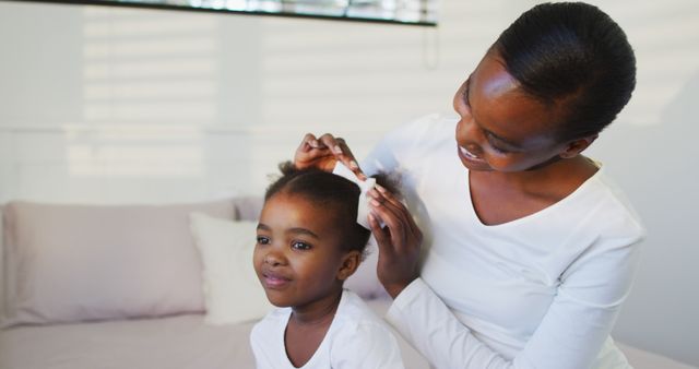A mother is styling her daughter’s hair in a bright, cozy bedroom. They are both smiling and enjoying the bonding moment, showcasing love and care between parent and child. This can be used for themes related to family, motherhood, parenting, haircare products, and lifestyle. Ideal for advertisements, blog posts, and social media content highlighting family values, home life, and children's daily routines.