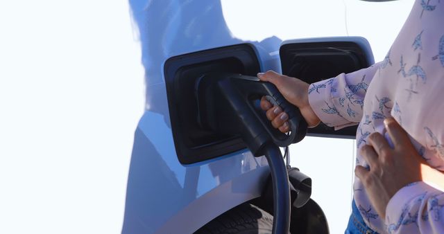 Person holds charging plug while connecting it to an electric vehicle outdoors on a bright day, emphasizing sustainable energy practices. Ideal for content related to green technology, eco-friendly transport, and renewable energy initiatives.