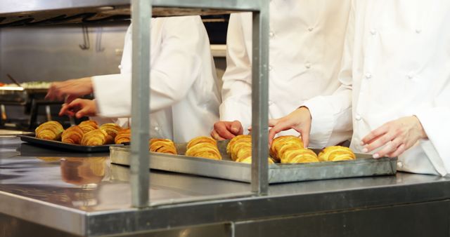 Chefs are baking croissants in a commercial kitchen. They are preparing pastries on trays, showcasing teamwork in a professional culinary environment. This could be used to depict gourmet cooking, culinary arts, teamwork in a commercial kitchen, or promotional materials for restaurants and baking schools.