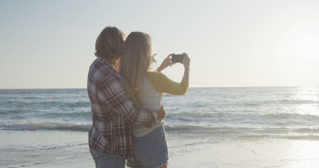 Couple stands side by side, taking a selfie with ocean in background during sunset. Useful for themes around love, romance, vacation, relaxation, and beach activities.