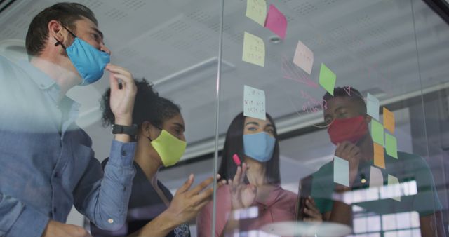 Diverse colleagues wearing face masks using memo notes on glass wall having a discussion. independent creative design business during covid 19 coronavirus pandemic.