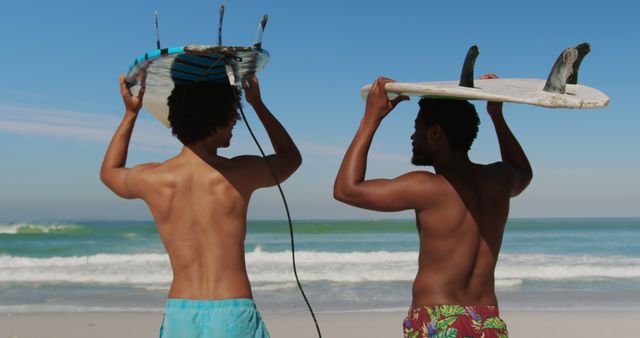 Two Afro male friends carrying surfboards on heads, preparing for surfing at beach, with ocean waves in background. Perfect for themes of friendship, adventure, summer activities, surfing lifestyle, beach vacations.