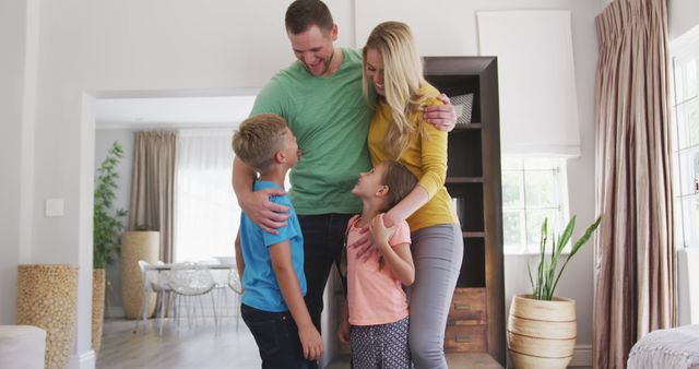 Parents hugging their two children in bright living room during daytime bonding. Perfect for use in advertising, family lifestyle, home, and parenting themes. Ideal for illustrating family values, affection, and joyful home atmosphere.