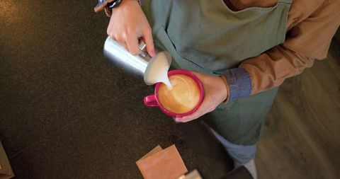 Female barista pouring steamed milk into red mug for latte art, cozy cafe counter
