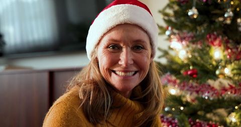 Mature Woman Smiling Wearing Santa Hat Near Christmas Tree Indoors