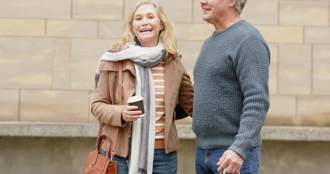 Senior couple smiling and holding coffee while strolling past tan stone wall in city
