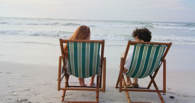 A Caucasian woman and a child are relaxing in beach chairs facing the ocean, with copy space. Their peaceful posture and the serene beach setting evoke a sense of tranquility and leisure.