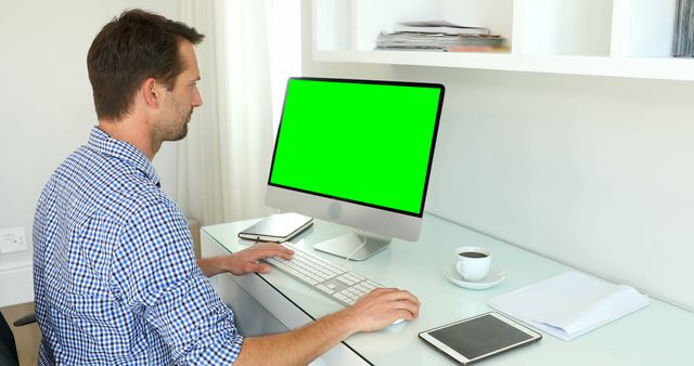 Man wearing checkered shirt working at clean, modern desk with a computer featuring a green screen display. Also on the desk are a tablet, documents, and a cup of coffee. Suitable for business, technology, and professional workspace themes. Useful for demonstrating tech apps, software interfaces, or remote working setups.