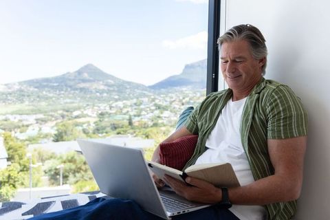 Mature man relaxing on window seat reading book and using laptop with mountain view