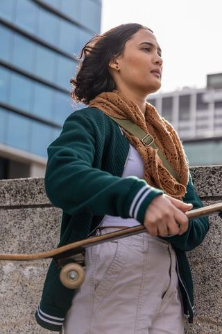 Asian woman leaning on concrete barrier holding skateboard wearing scarf in urban plaza