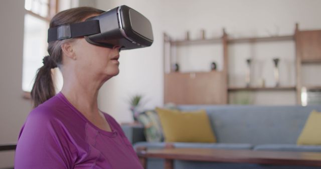 Woman wearing VR headset sitting in modern living room. This image is great for illustrating advanced technology, gaming, interactive digital experiences, seniors engaging with modern tech, or home relaxation with immersive devices.