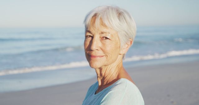 Senior woman with short gray hair smiling by the beach. Ideal for illustrating themes of retirement, happiness in old age, coastal living, leisure activities, and enjoying nature. Useful for advertisements, magazine articles, blogs, and wellness content targeting senior demographics.