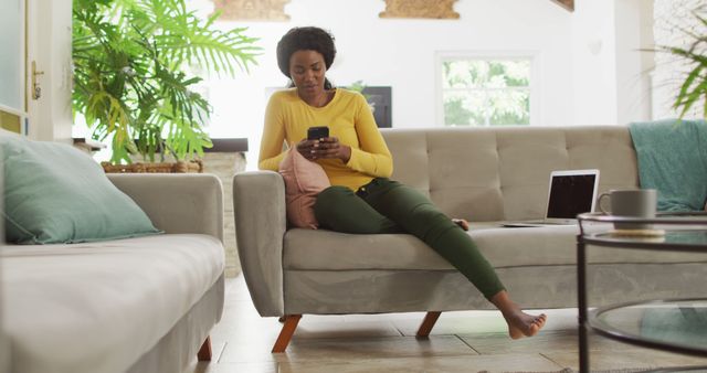 Woman in casual clothing lounging on a comfortable sofa while checking her smartphone. Nearby, there is an open laptop, indicating a multi-use space. Bright and airy living room with cozy cushions and green plants adding a fresh touch. Ideal for themes related to home life, technology use, modern living, remote work, leisure activities, or casual relaxation.