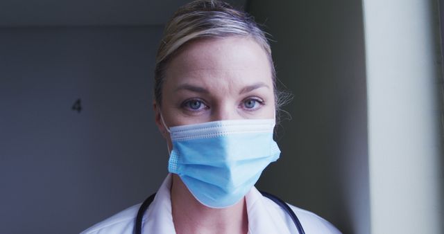 Female doctor standing indoors wearing a protective medical mask and stethoscope, expressing seriousness and professionalism. Can be used for healthcare, medical services, pandemic response, or safety protocol related content.