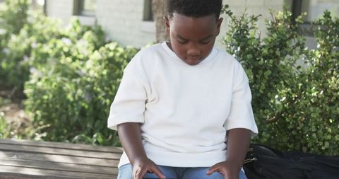Young boy sitting on wooden bench looking at lap with backpack in landscaped garden