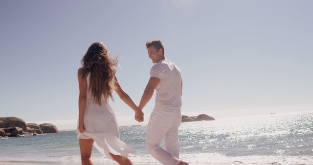 A young Caucasian couple enjoys a romantic walk along a sunny beach, with copy space. Their carefree stroll by the sea captures a moment of love and connection.