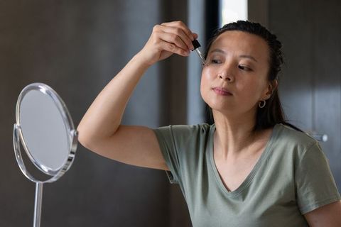 Mature asian woman applying facial serum with dropper at vanity mirror, skincare routine