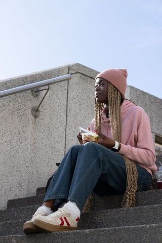African american woman sitting on steps holding salad, wearing pink hoodie and beanie