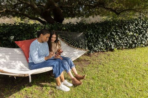 Two women relaxing on hammock sharing smartphone and takeout coffee in sunny garden