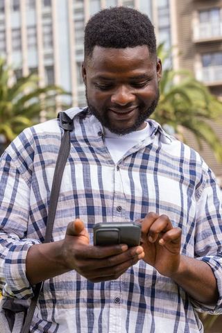 Smiling african man using smartphone in urban plaza wearing plaid shirt and shoulder bag