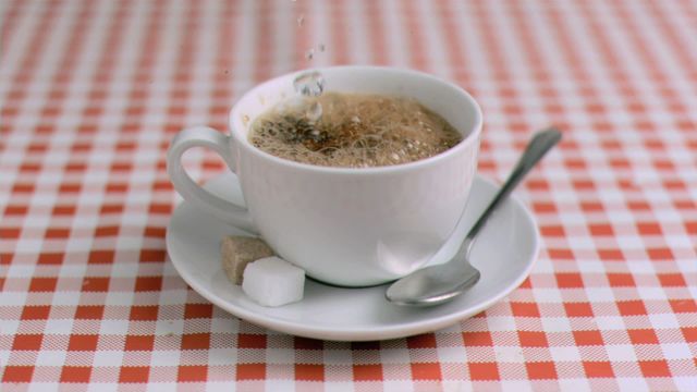 Water dripping in super slow motion into a cup of instant coffee placed on a white saucer with a spoon and sugar cubes, all on a red and white checkered tablecloth. Ideal for use in content related to breakfast, coffee culture, beverages, slow-motion effects, and food blogs.