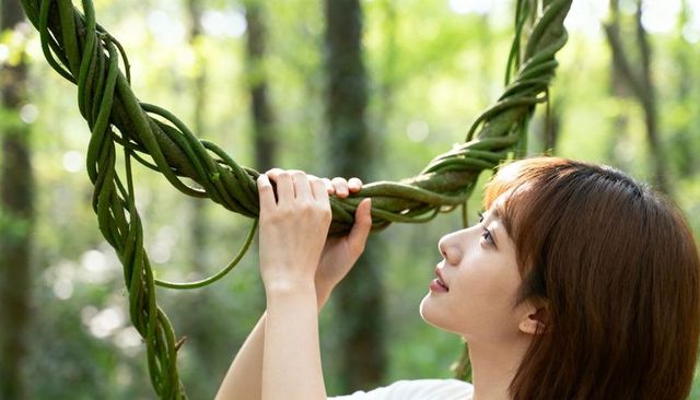 Asian woman reaching and grasping twisted vine loop in sunlit green forest, serene profile portrait