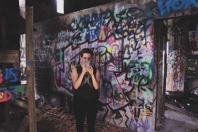 Young woman capturing a photo on her phone while exploring an abandoned building with graffiti covered walls. Ideal for promoting urban exploration, street art culture, or photography adventurers. Use in blogs, social media, and artistic projects highlighting urban decay and hidden city art.