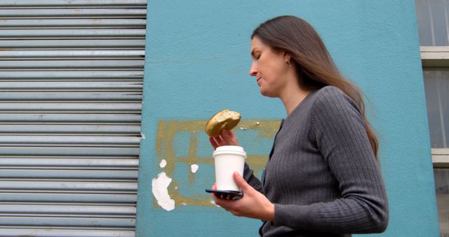 Woman eating donuts while walking on street. Woman holding coffee cup and mobile phone 4k